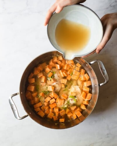 a woman pouring broth into a stainless steel pot of chicken, celery, cubed sweet potatoes, and seasoning