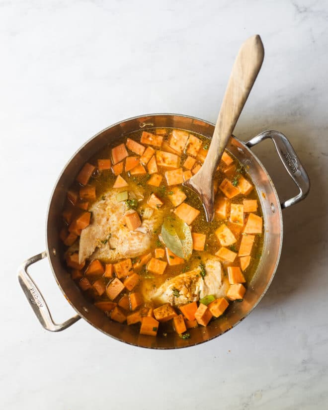 a pot of broth, cubed sweet potatoes and chicken breast with a wooden spoon sticking out of it