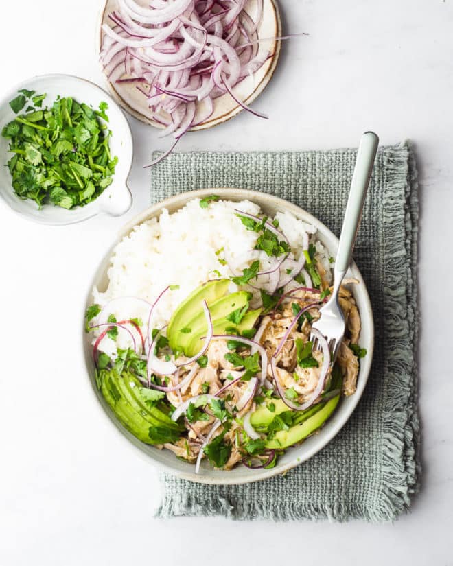 a bowl of salsa chicken and rice garnished with cilantro, avocado, limes, and red onion