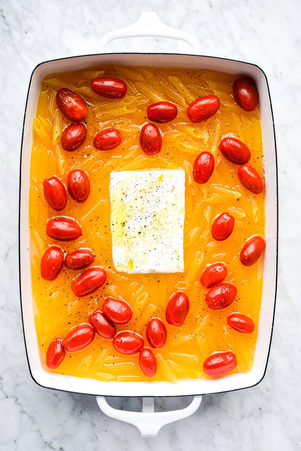 a large white baking dish filled with uncooked penne pasta, broth, cherry tomatoes, and a feta cheese block in the middle