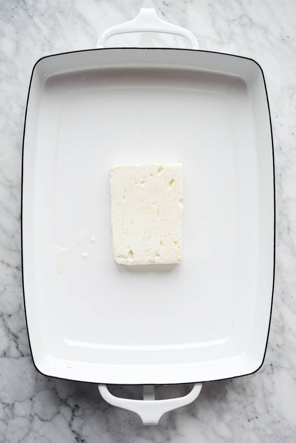 a block of feta cheese sitting in a white baking dish on a marble surface