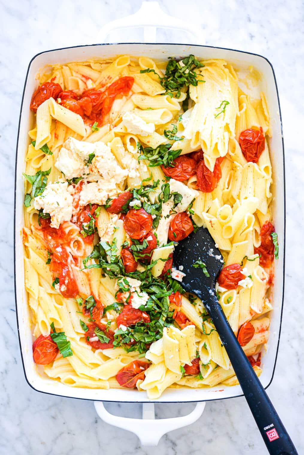 a white baking dish with pasta, cherry tomatoes, feta, garlic, and basil, being stirred up with a black rubber spatula