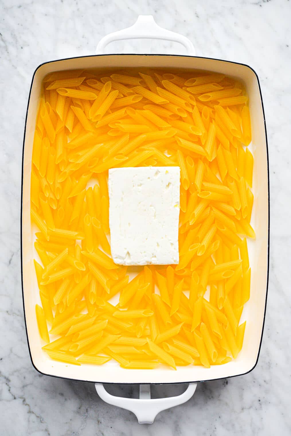 a white baking dish with uncooked penne pasta and a block of feta cheese in it