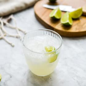 a salt rimmed glass of keto margarita sitting in front of a cutting board with lime juice wedges sitting on it