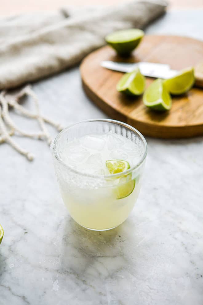 a salt rimmed glass of keto margarita sitting in front of a cutting board with lime juice wedges sitting on it