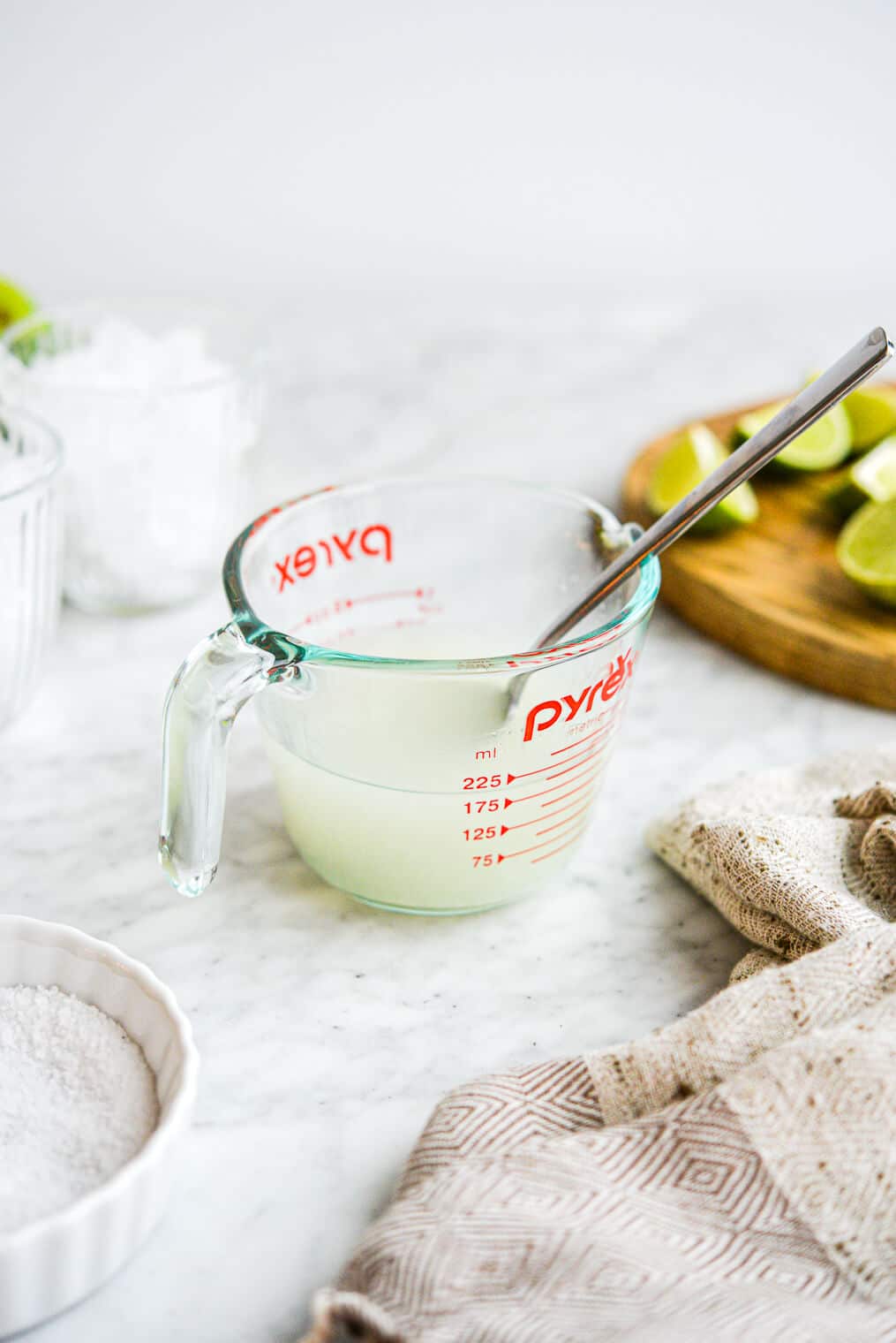 a measuring cup of keto margarita sitting in front of glasses of ice, a cutting board of lime wedges, and sitting behind a small bowl of salt