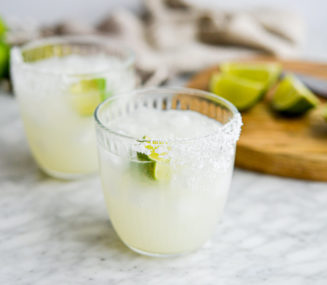 two salt rimmed glasses of keto margarita sitting in front of a cutting board with lime juice wedges sitting on it
