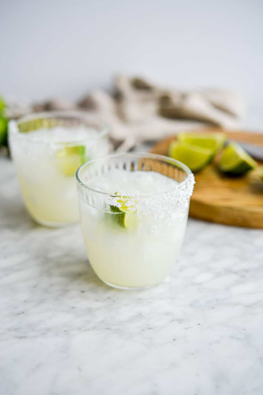 two salt rimmed glasses of keto margarita sitting in front of a cutting board with lime juice wedges sitting on it