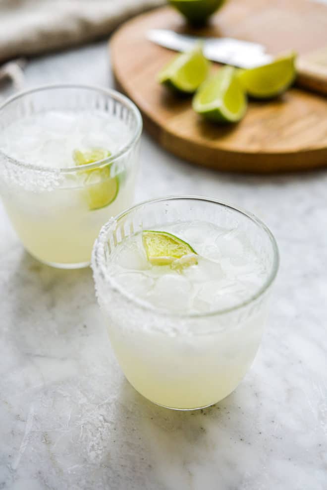 two salt rimmed glasses of keto margarita sitting in front of a cutting board with lime juice wedges sitting on it