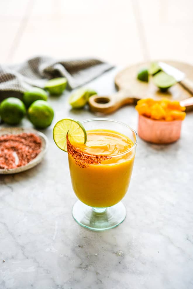 a chili salt rimmed glass of frozen mango margarita with the ingredients for mango margaritas in the background