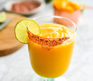a chili salt rimmed glass of frozen mango margarita with the ingredients for mango margaritas in the background