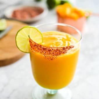 a chili salt rimmed glass of frozen mango margarita with the ingredients for mango margaritas in the background