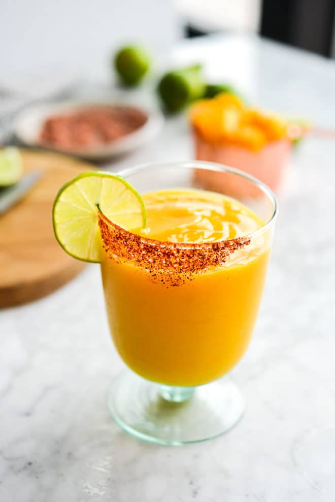 a chili salt rimmed glass of frozen mango margarita with the ingredients for mango margaritas in the background