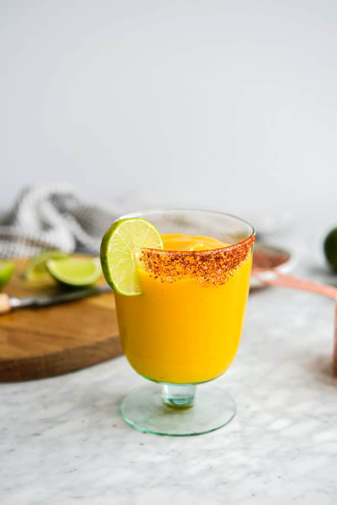 a chili salt rimmed glass of frozen mango margarita with the ingredients for mango margaritas in the background