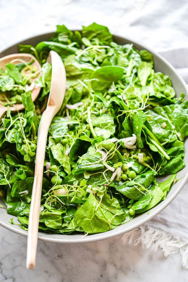 a large gray bowl filled with a bright green spinach salad with two salad spoons sticking out of the bowl