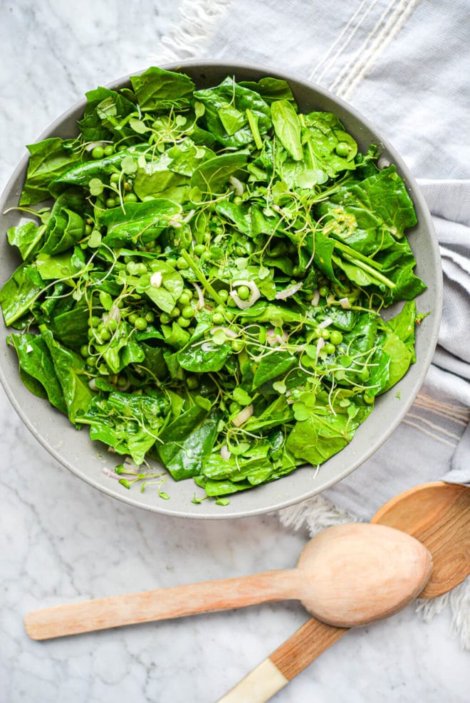 a large gray bowl filled with a bright green spinach salad with two salad spoons sticking out of the bowl