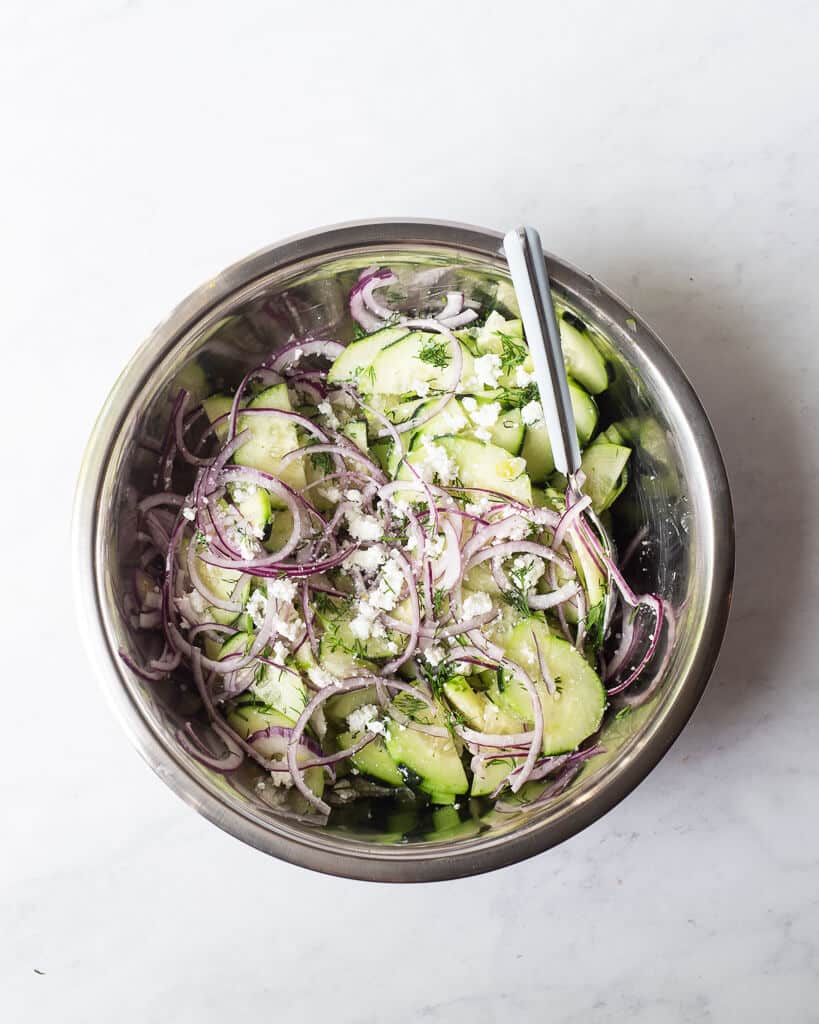 a greek cucumber salad made of thinly sliced cucumber, red onion, feta cheese, and dill in a large metal mixing bowl