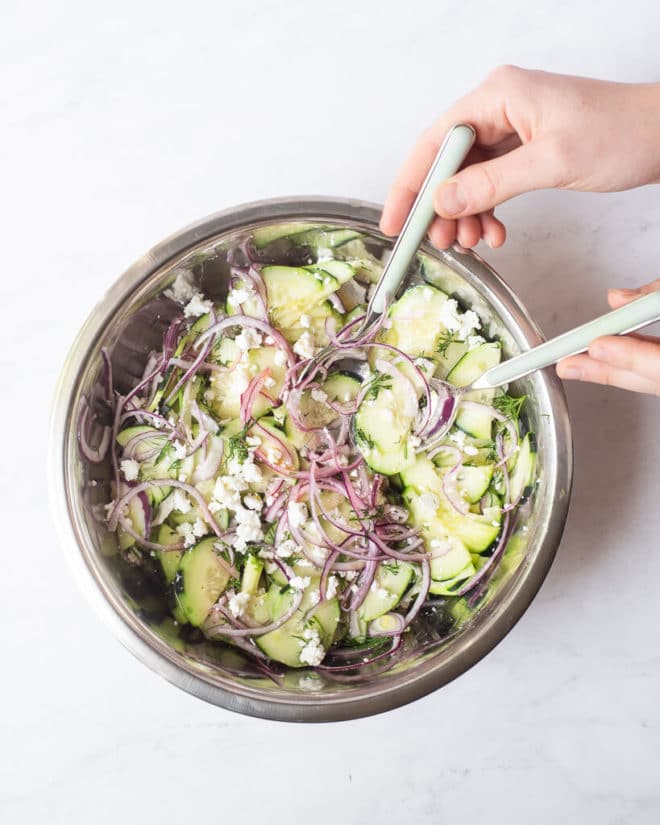 a greek cucumber salad made of thinly sliced cucumber, red onion, feta cheese, and dill in a large metal mixing bowl with a person's hands mixing everything together with two forks