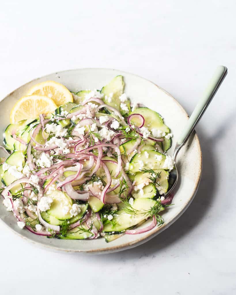 a greek cucumber salad made of thinly sliced cucumber, red onion, feta cheese, and dill sitting on a light gray plate