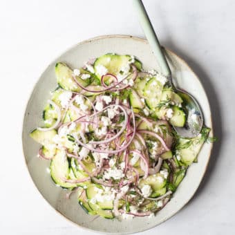 a greek cucumber salad made of thinly sliced cucumber, red onion, feta cheese, and dill sitting on a light gray plate