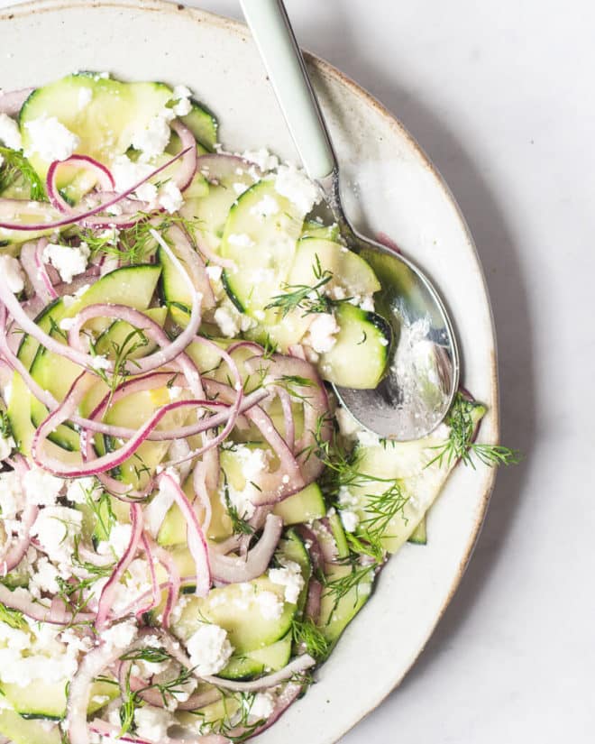 a greek cucumber salad made of thinly sliced cucumber, red onion, feta cheese, and dill sitting on a light gray plate