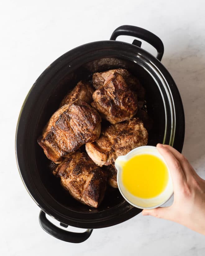 seared pieces of pork shoulder in a slow cooker with a person's hand pouring a small bowl of orange juice over top