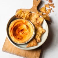 a wooden cutting board with a bowl of vegan nacho cheese and hand fried corn tortilla chips sitting on top of it
