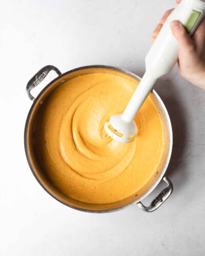 a large pot of vegan nacho cheese with a person holding an immersion blender over it