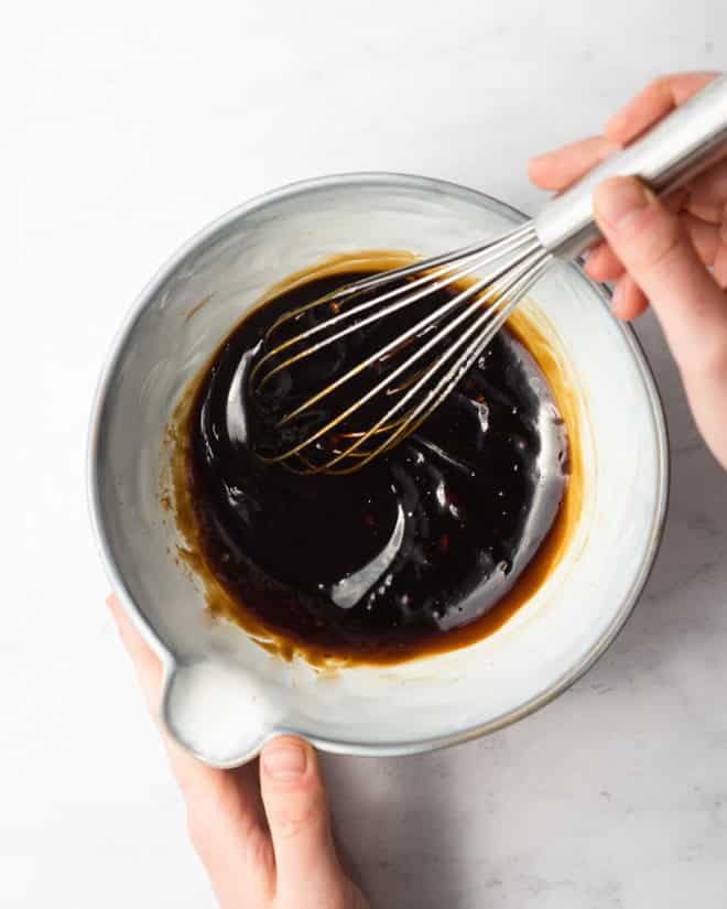 woman's hands whisking a bowl of teriyaki sauce in a grey bowl on a marble surface