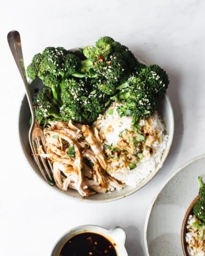 teriyaki chicken bowl with white rice and broccoli garnished with sesame seeds and red pepper flakes on a marble surface