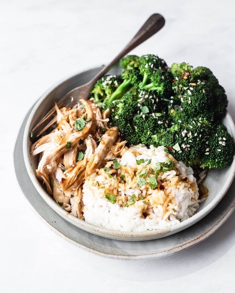 side view of a teriyaki chicken bowl filled with shredded chicken, broccoli florets, and white rice drizzled with teriyaki sauce, sesame seeds, and red pepper flakes