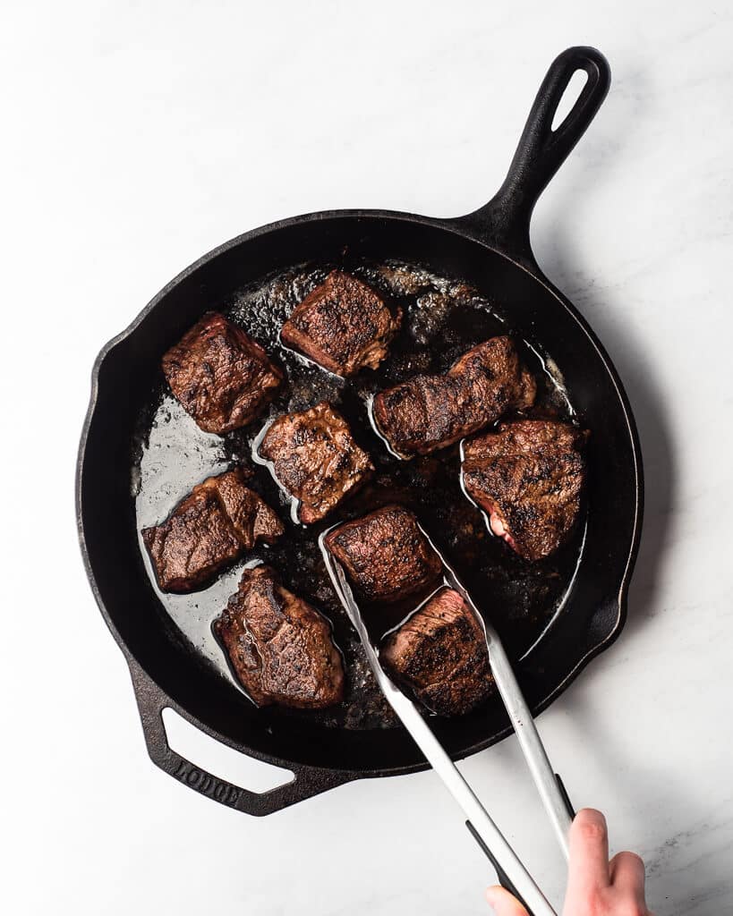 cast iron skillet on a marble surface filled with large chunks of seared beef with one being held by a pair of tongs