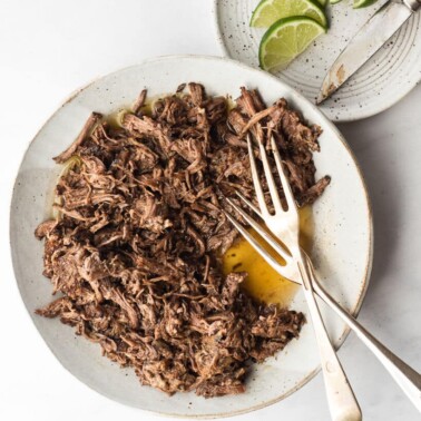 a grey plate filled with shredded barbacoa and two forks next to a white speckled plate with lime wedges and a knife on a marble surface