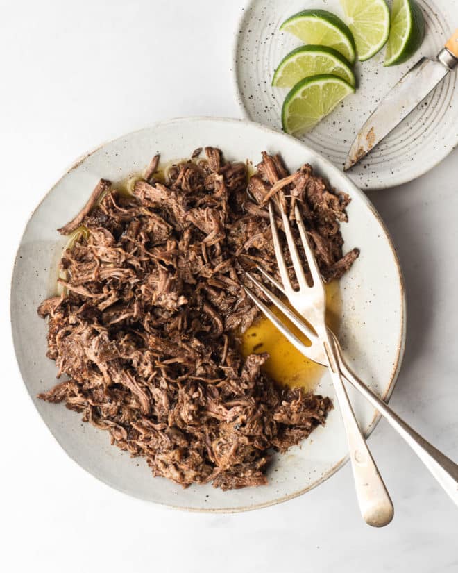a grey plate filled with shredded barbacoa and two forks next to a white speckled plate with lime wedges and a knife on a marble surface