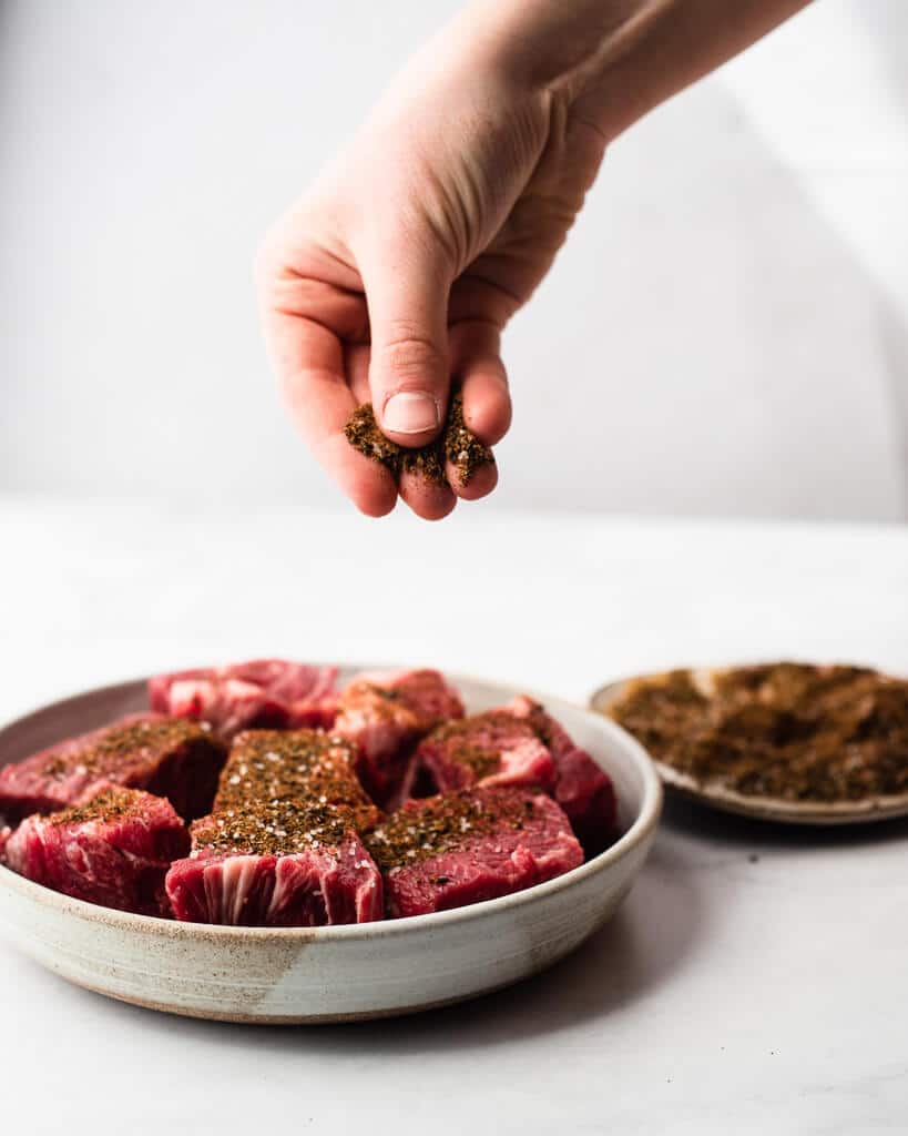 woman's hand seasoning a plate full of chunks of bright red beef