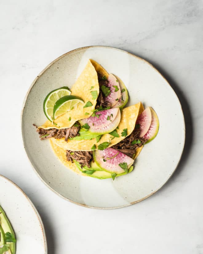 grey plate with two corn tortilla tacos filled with barbacoa and topped with sliced avocado, watermelon radishes, chopped cilantro, and lime wedges on a mabrle surface