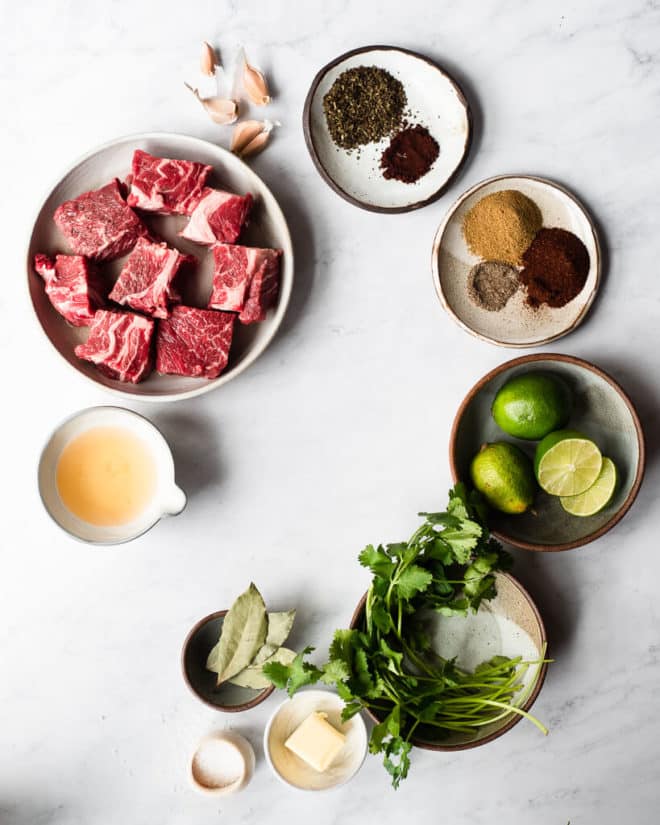 plates with barbacoa taco ingredients on a marble surface including chunks of chuck roast, garlic cloves, spices, limes, cilantro, bay leaves, salt, butter, and apple cider vinegar
