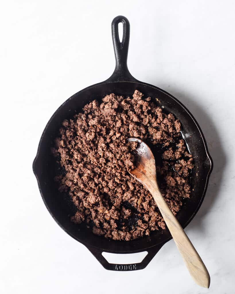 a person holding a wooden spoon to brown ground meat in a cast iron skillet