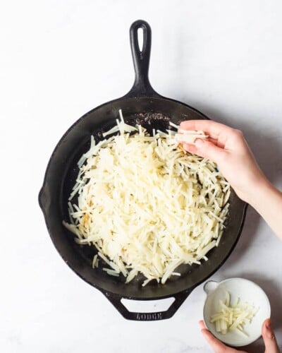a person adding shredded hash browns to a cast iron skillet