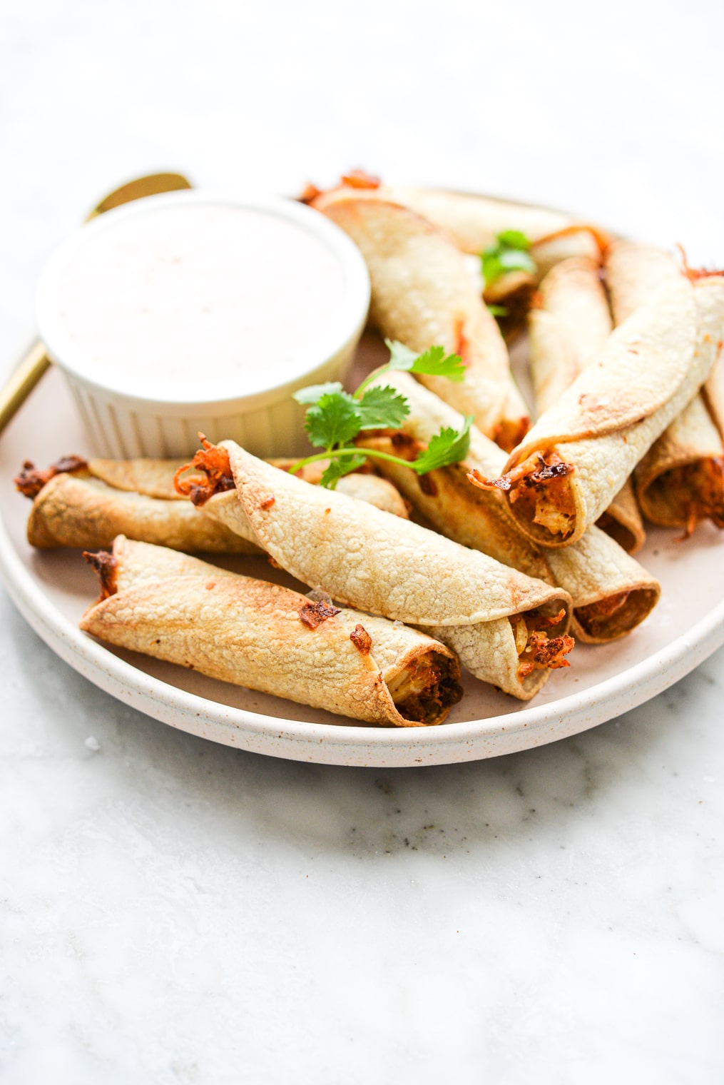 a plate of air fryer chicken taquitos and a small bowl of sour cream sitting on a marble surface