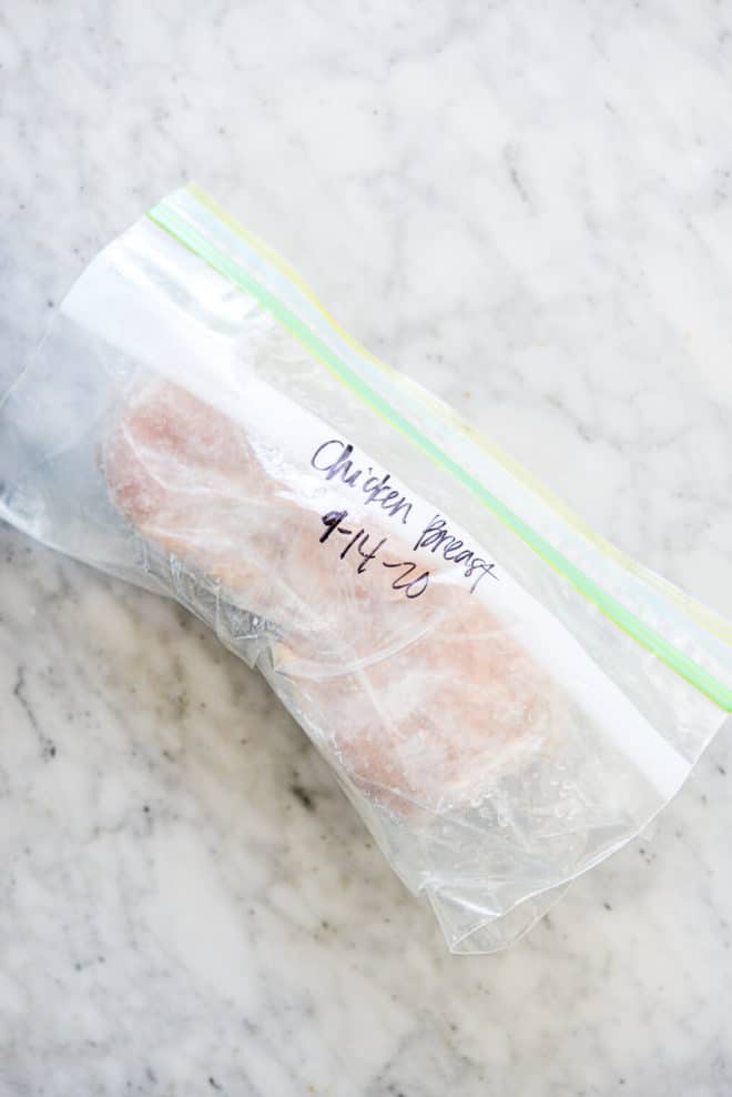 a ziplock bag labeled "chicken breast 9-14-20" with frozen chicken breast inside of it sitting on a marble surface