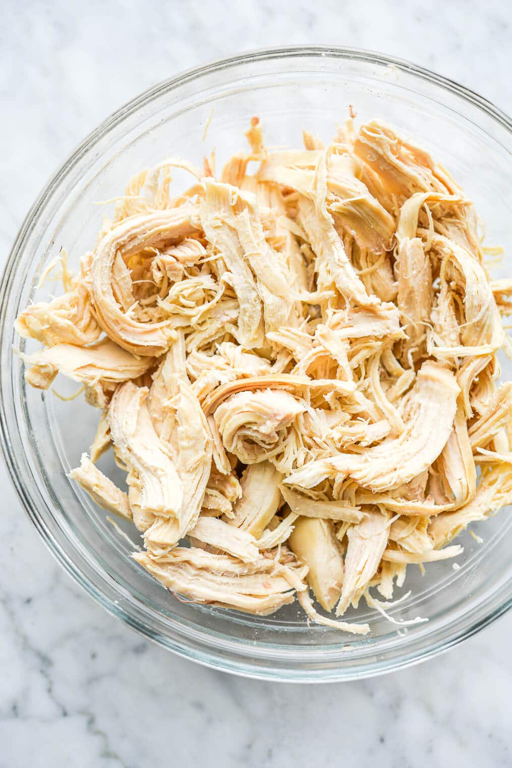 a clear glass bowl of shredded chicken sitting on a marble surface