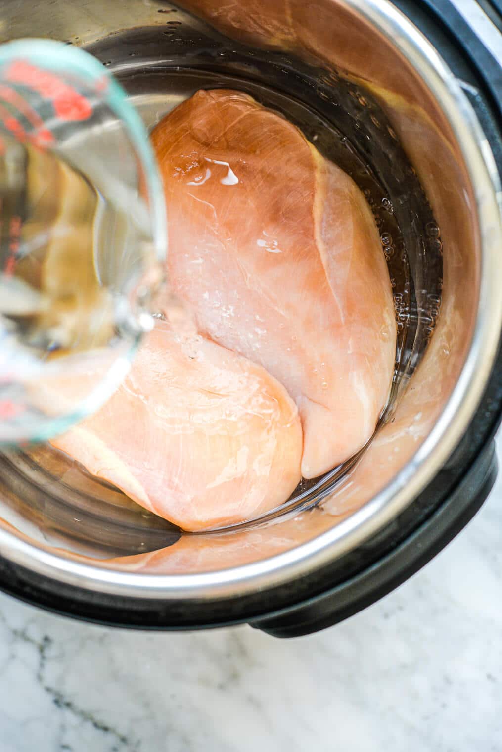 a person pouring a cup of water over two raw chicken breasts in an instant pot