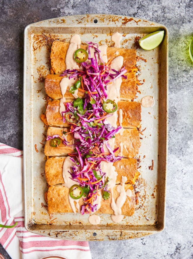 chicken chimichangas topped with a creamy light orange sauce and a bright purple cabbage slaw on a sheet pan on a dark grey surface