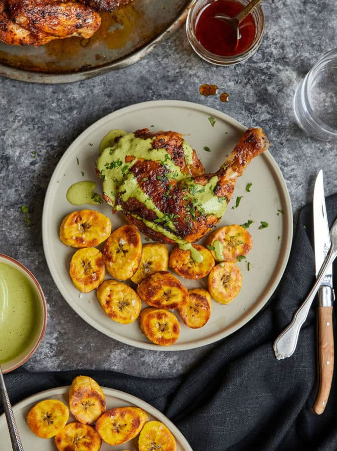 chicken leg quarter with crispy skin on a grey plate surrounded by sliced fried plantains and drizzled with creamy green sauce on a dark grey surface