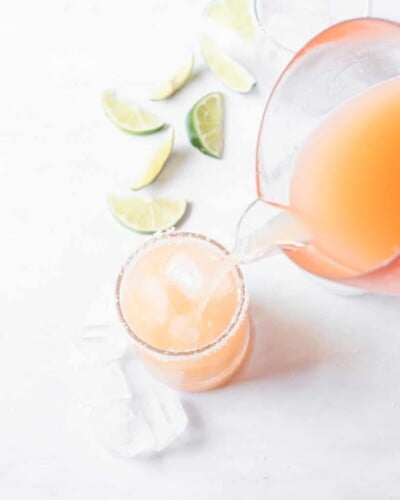 a pitcher of palomas being poured into a salt rimmed glass with cut limes in the background