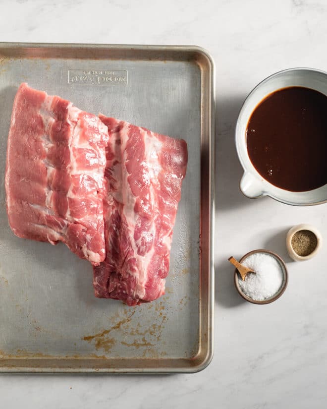 two raw racks of baby back ribs on a sheet pan next to small bowls of salt and pepper and a larger bowl of BBQ sauce