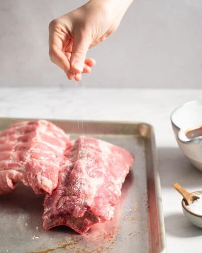 a person seasoning raw racks of baby back ribs on a rimmed sheet pan