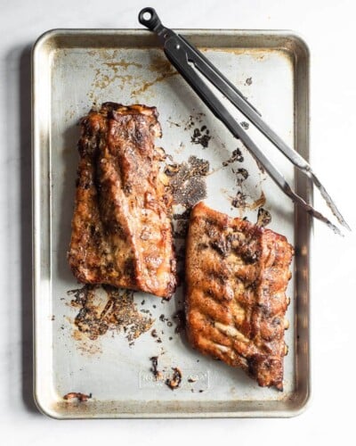 two half racks of rubs without BBQ sauce sitting on a rimmed baking sheet next to a pair of kitchen tongs