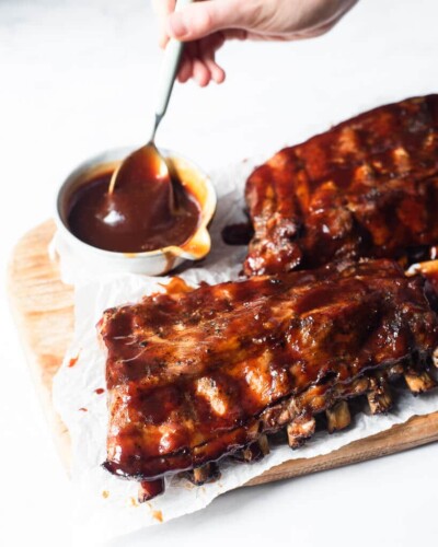 two half racks of ribs smothered in BBQ sauce sitting on a wooden cutting board next to a bowl of more BBQ sauce